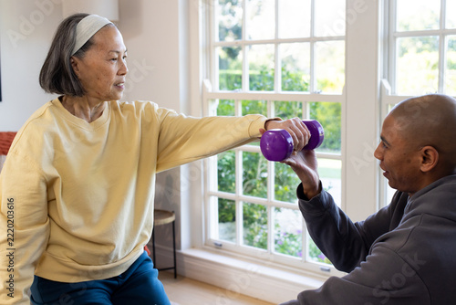 Diverse coach kneeling helping Senior Asian woman in sweatshirt raising purple dumbbell at home
