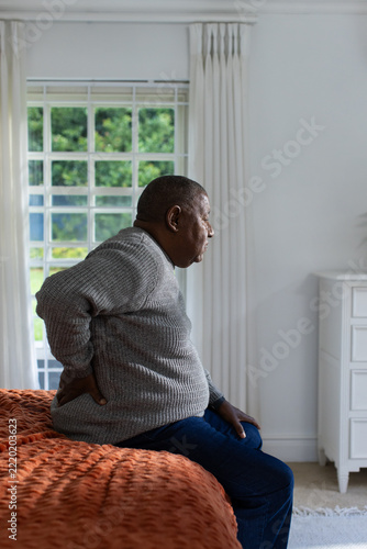 Senior African American man sitting on bed in bedroom holding back wearing sweater rust coverlet