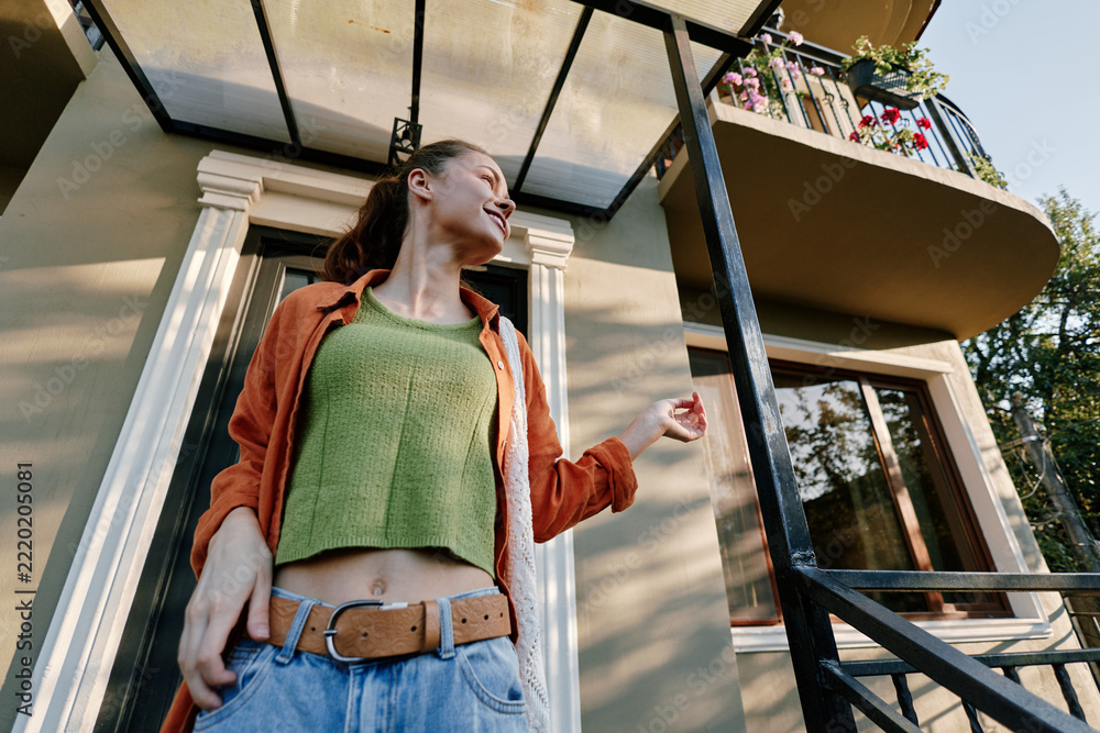 Naklejka premium Young woman wearing a green knit sweater and orange jacket stands outside a modern home, smiling confidently. Casual fashion concept for lifestyle and streetwear.