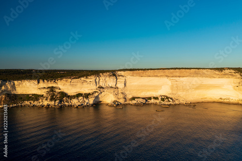 Limestone cliffs line the coast of Bonifacio above calm water and under a clear blue sky. Golden light highlights the rock layers and shoreline.