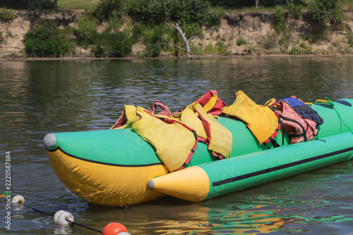 Green and yellow inflatable banana boat with colorful life jackets floating beside a tree-lined riverbank