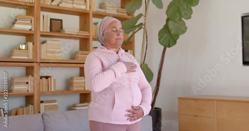 Senior African American woman standing eyes-closed pink top hands on chest belly breathing at home