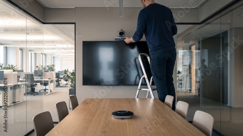 IT Technician Installing Video Conference Camera Above Wall Mounted Display in Modern Corporate Meeting Room with Conference Table and Audiovisual Equipment