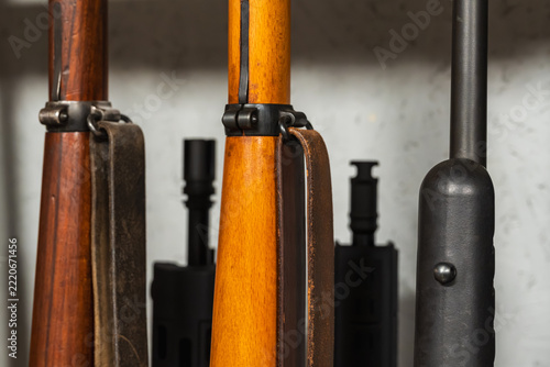 Close up view of vintage wooden stocks with leather straps alongside modern metal rifle barrels stored in armory vault gun rack cabinet