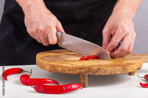 Close-up view of the hands of a male cook who cuts red chili peppers with a knife on a cutting board, selective focus. Episode of the cooking process