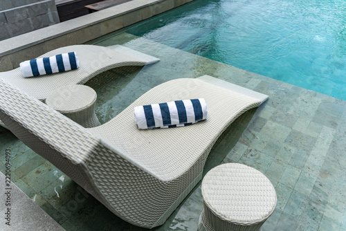 A high-angle or eye-level shot featuring a crisp blue and white towel lying on a modern sun lounger by a clear, sparkling turquoise swimming pool.
