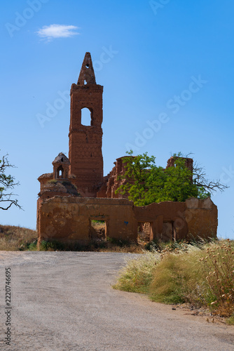 Weathered adobe-style church ruins rise above a quiet country lane under a bright, cloudless sky. An ornate brick tower, empty doorways and a lone leafy tree show the decay of this forgotten structure