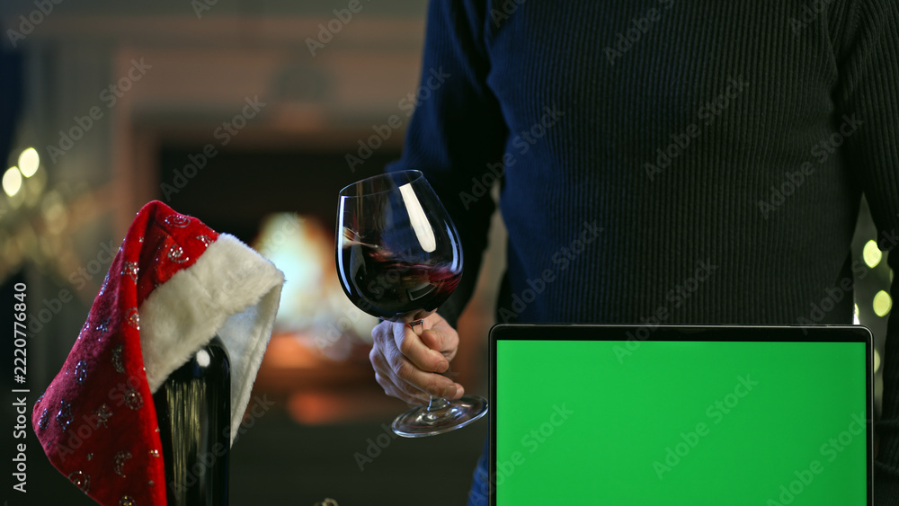 Naklejka premium Glass and bottle of red wine with laptop computer on table in Christmas decorated home. Man tasting buying wine online. Green screen for web shop ads and vinery holiday offer.