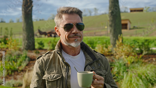 Mature age rancher drinking cup of coffee on sunny day, with horses in the background. Landowner enjoying successful agricultural business on ranch. Mid adult man with gray hair smiling outdoor.