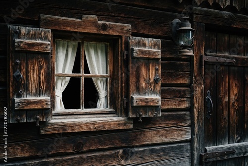 Old wooden window with open shutters, white curtains on dark rustic cabin wall. Vintage style house exterior features rustic wood texture, antique lantern. Cozy mountain chalet architecture in winter