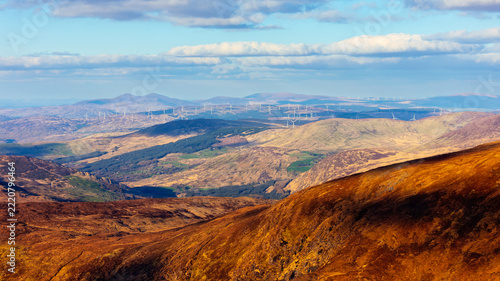 An aerial view of a mountainous landscape with rolling hills, distant wind turbines, and a blue sky with clouds.