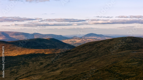 Golden afternoon light illuminates the vast grassy slopes and distant mountain ranges in Ireland as soft clouds drift across the horizon during the daytime.