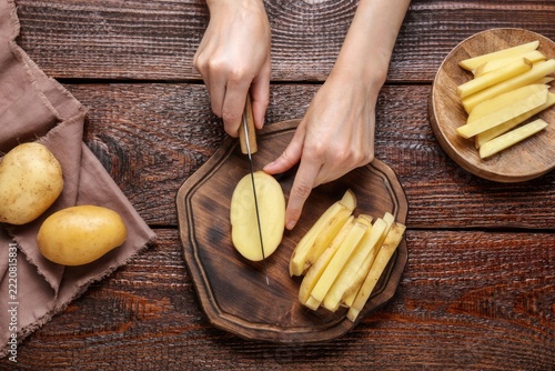 Preparing Fresh Potato Fries: Hands skillfully slice raw potatoes on a wooden board, surrounded by whole potatoes and cut fries, ready to be transformed into a delicious snack.