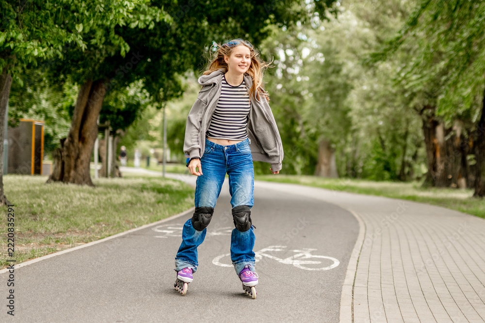 Fototapeta premium Happy young girl rollerblades on paved park path. Green trees line route. Sunny daytime activity.