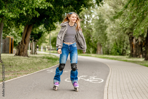 Happy young girl rollerblades on paved park path. Green trees line route. Sunny daytime activity.