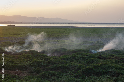 Smoke Rising From Grassland Near Lake at Sunset
