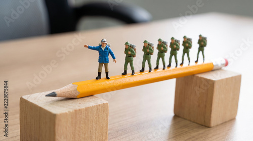 Miniature team leader guiding line of figurines across pencil bridge balanced on wooden blocks, leadership and teamwork concept