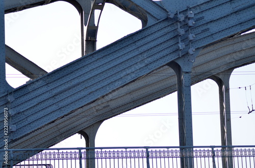 An industrial steel bridge construction showing a close-up of complex metallic geometric beams and rivets against a clear blue sky background. Ideal for engineering and architecture concepts.