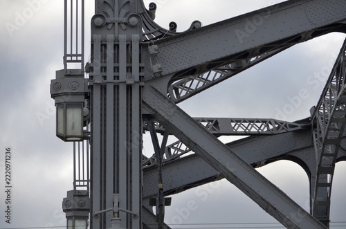 An industrial steel bridge construction showing a close-up of complex metallic geometric beams and rivets against a clear blue sky background. Ideal for engineering and architecture concepts.