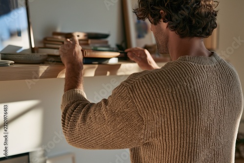 Young man arranging books on wooden shelf in warm sunlit minimalist home interior