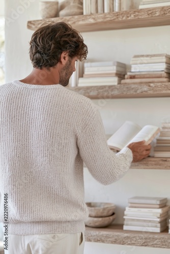 Young man reading open book beside rustic wooden shelves in bright minimalist home library