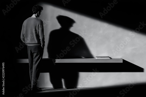 Man standing beside minimalist shelf with book and dramatic shadow in black and white interior