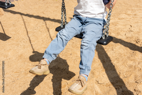 Child sitting on a swing at a sandy playground wearing blue joggers and tan sneakers