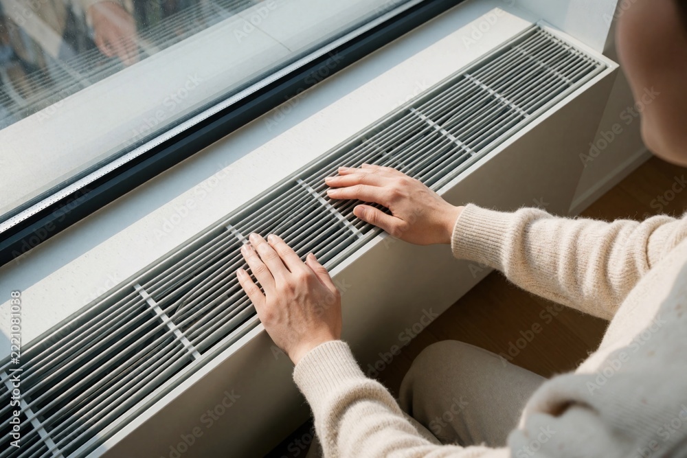 Fototapeta premium Person adjusts modern white radiator near window in home. Hand touches heater grill to control indoor temperature. Man manages heating energy, utility costs during winter. Efficient thermal