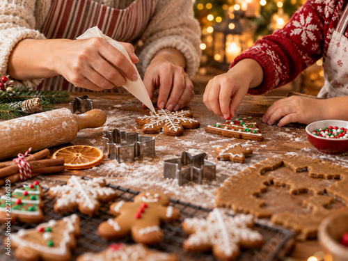 Close-up of mother and child decorating homemade Christmas gingerbread cookies with white icing and colorful sprinkles on a wooden kitchen table. Festive baking scene with snowflake and Christmas tree