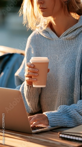 A faceless young caucasian student sits on a wooden bench outdoors while holding a coffee cup and tilting her head towards an open laptop for study.