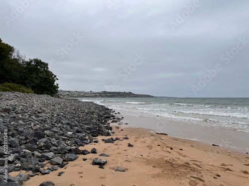 A sandy beach with dark rocks and pebbles along the left edge meets gently breaking waves under an overcast sky, with a wooded bank on the left and a headland with buildings in the distance.
