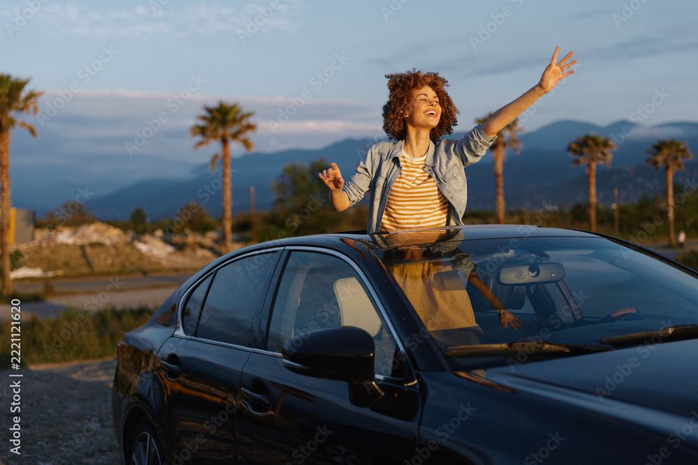 Fototapeta premium A woman leans from a sleek car, arms raised in joy, wearing a striped shirt and denim jacket. The sunny outdoors with palm trees and distant mountains conveys freedom and lifestyle.