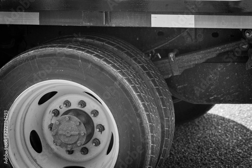 Black and white close-up photograph of a heavy-duty vehicle tire and wheel beneath a truck chassis. The image highlights the tire tread pattern, metal wheel rim, lug nuts, central hub, and suspension 