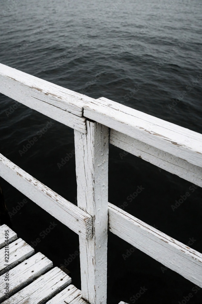 Fototapeta premium White wooden fence and pier railing over dark ocean water. Old weathered wood structure stands near sea. Close up view of maritime construction on coastal boardwalk during summer.