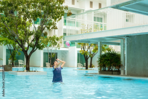 Happy Asian girl playing with a ball in a swimming pool at a tropical white resort, young person enjoying summer vacation, leisure and outdoor recreation. Fun childhood lifestyle and holiday concept.
