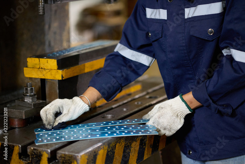Worker wearing a blue uniform and white gloves meticulously inspects a metal component with small holes in a factory