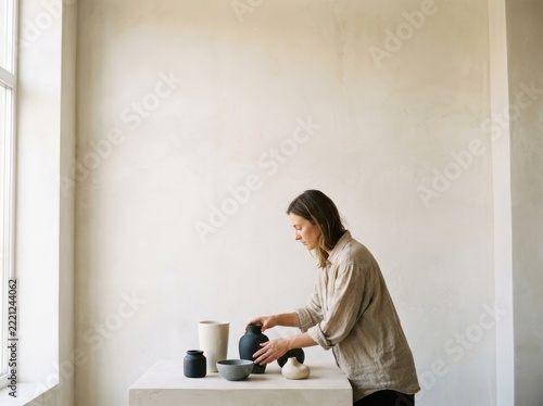 A woman arranges ceramic objects on a white table near a window. The setting is minimalist with a plain wall in the background. The woman is wearing a light-colored shirt and appears focused
