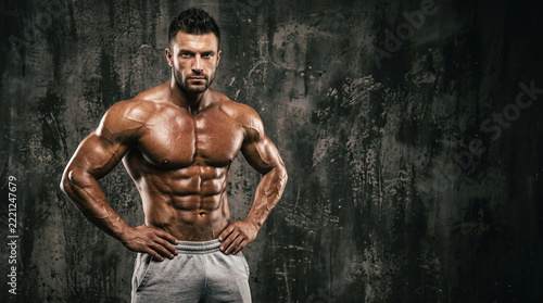 Strong muscular bodybuilder standing with hands on hips against dark grunge wall, shirtless fitness model showing athletic physique and six pack abs with copy space for workout motivation.