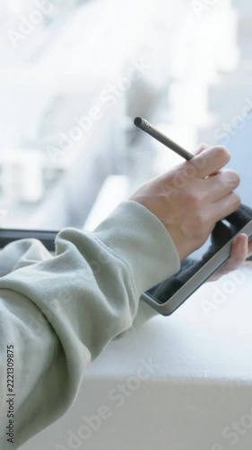 Vertical video: Lowering stylus starting person sketching on tablet at window ledge in sage top