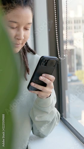 Vertical video: Sitting adult with locs, pale tee reading phone at sill, leaf shifting