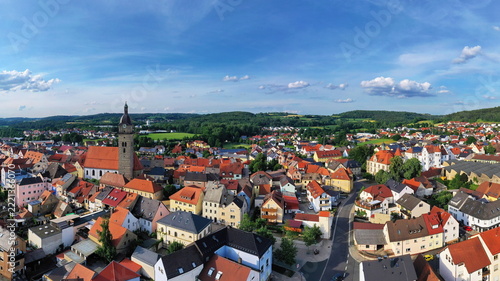 Malerische Luftaufnahme von Auerbach in der Oberpfalz mit der markanten Stadtpfarrkirche, traditionellen roten Dächern und idyllisch grüner Landschaft im Hintergrund unter blauem Himmel.