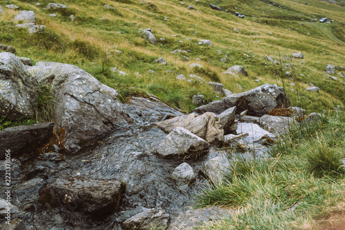 Mountain stream. Water flows between large rocks surrounded by green and golden grasses on a mountain hillside.