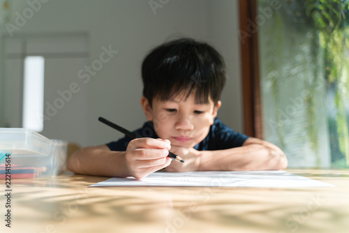 Young Asian Boy Writing with Pen at Table by Window in Natural Light