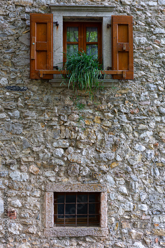 Rustic stone house facade with wooden shuttered window and green plant in the medieval village of Canale di Tenno, Trentino, Italy.