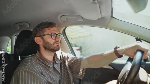 Young man driving car on sunny day, focused male driver with glasses holding steering wheel while commuting through green countryside on summer road trip