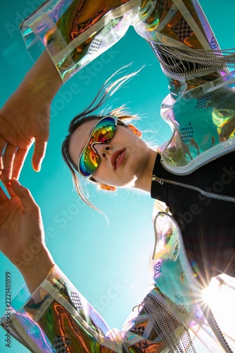 Model poses under bright sky wearing reflective sunglasses and a transparent jacket, capturing a moment of modern fashion in outdoor light