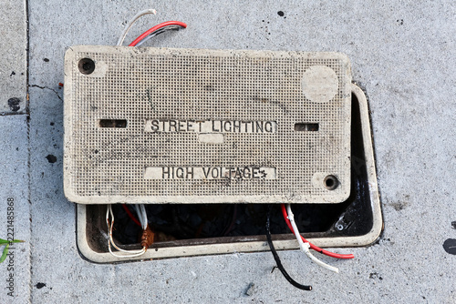 Open electrical utility vault with exposed cables beneath a high voltage street lighting warning on the sidewalk