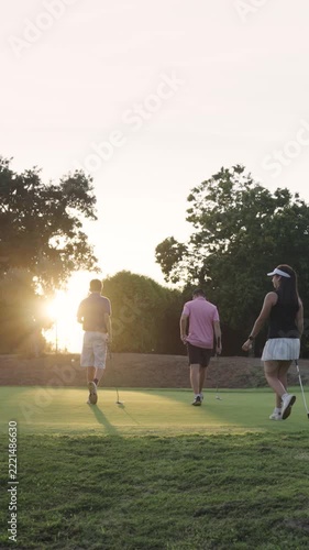Group of young golfers walking on a beautiful golf course. Bright sun shines behind them as they carry their putters across the green. Vertical
