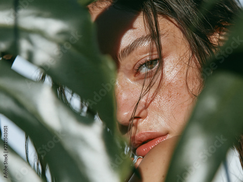 A freckled young woman with wavy brown hair in burnt‑orange garment gazes at viewer, framed by lush large green tropical plant leaves in soft natural light.