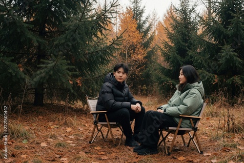 Young Asian couple sits on camping chairs in autumn forest. Man, woman enjoy peaceful outdoor recreation under pine trees. Lovers relax together during fall vacation trip in woods, feeling calm,